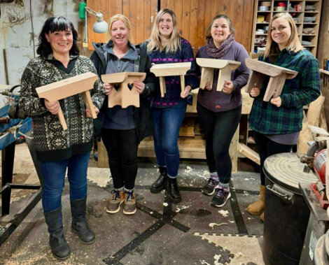 Five women stand indoors holding small wooden stools they built; the space appears to be a woodworking workshop with tools and wood pieces visible.