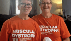 Two smiling people wearing orange "Muscular Dystrophy UK, Our Muscles Matter" shirts stand indoors, holding a skein of white yarn.