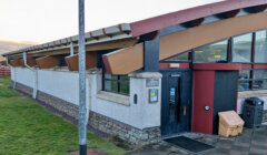Single-story modern building with a curved roof, stone and white walls, a black door, and a ramp; grass and a lamppost in the foreground.
