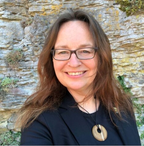 Woman with long brown hair and glasses smiles at the camera, wearing a black top and a round pendant, standing in front of a rocky stone wall.