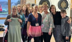 A group of women and one boy pose indoors; some wear lanyards, one woman holds a dress, and another wears a pink tutu. Dartboards and a table are visible in the background.