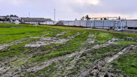 A muddy sports field with tire tracks and patches of worn grass; goalposts and buildings are visible in the background under an overcast sky.