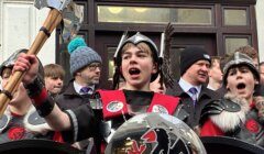 A group of people wearing Viking costumes, with one boy in the center holding an axe and shield, standing in front of a building.