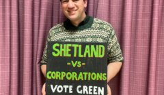 A person stands in front of a purple curtain holding a sign that reads, "SHETLAND vs CORPORATIONS VOTE GREEN.