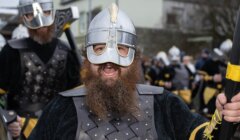 A person dressed as a Viking with a helmet and armor shouts during a reenactment event, surrounded by others in similar costumes.
