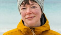 A person wearing a yellow jacket and gray knitted headband stands on a beach with the ocean and cloudy sky in the background.