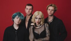 Four people pose against a solid red background, wearing black clothing and accessories, with varied hairstyles including blue, blonde, and short brown hair.