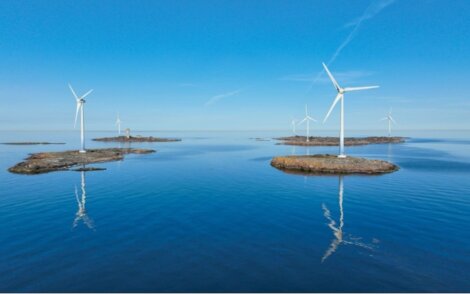 Wind turbines are installed on small rocky islands in a calm blue sea under a clear sky, with reflections visible on the water.