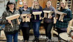 Five women stand indoors in a workshop, each holding a small wooden stool they have made. Various woodworking tools and materials are visible in the background.