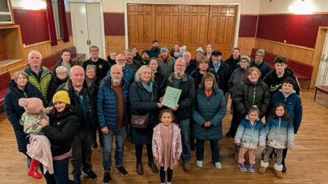 A group of people of various ages stand together indoors, some smiling, with one person in the centre holding a framed document or plaque.