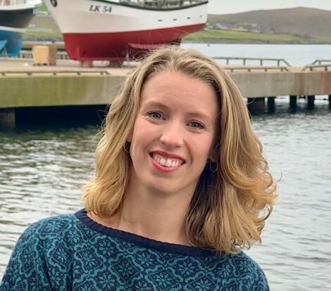 A woman with shoulder-length blonde hair smiles by a waterfront with a dock, boats, and hills in the background.