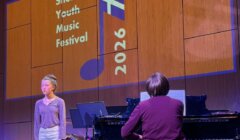 A girl stands singing beside a seated pianist on stage at the Shetland Youth Music Festival 2026, with the event’s logo projected on a wooden background behind them.