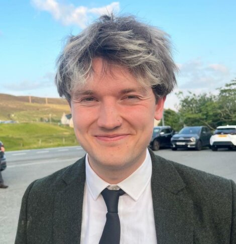 A person with short, tousled dark and grey hair, wearing a suit and tie, stands outdoors near a road with parked cars and grassy hills in the background.
