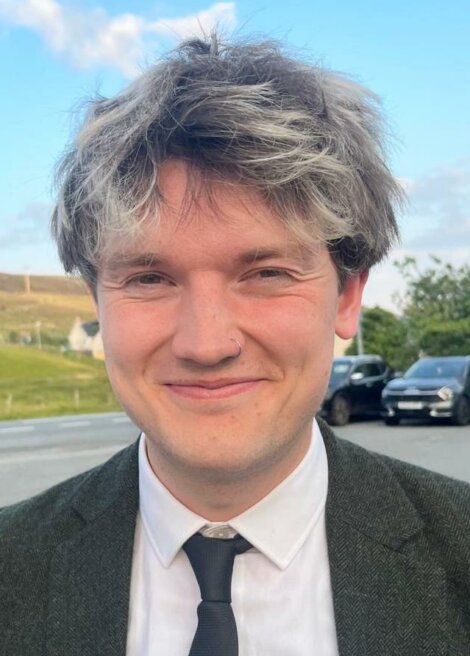 A person with short, tousled dark and grey hair, wearing a suit and tie, stands outdoors near a road with parked cars and grassy hills in the background.