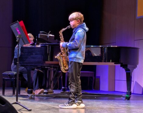 A boy in a blue hoodie plays an alto saxophone on stage, accompanied by a pianist seated at a grand piano.