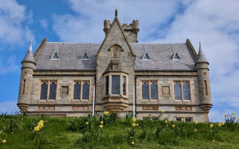 A stone Gothic-style building with pointed roofs and arched windows stands behind a grassy hill dotted with yellow daffodils under a partly cloudy sky.