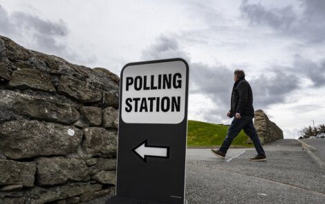 A sign reading "Polling Station" with an arrow points left as a person walks on a path beside a stone wall under a cloudy sky.