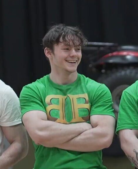 A young man with brown hair, wearing a green T-shirt with yellow lettering, stands with arms crossed and smiles.