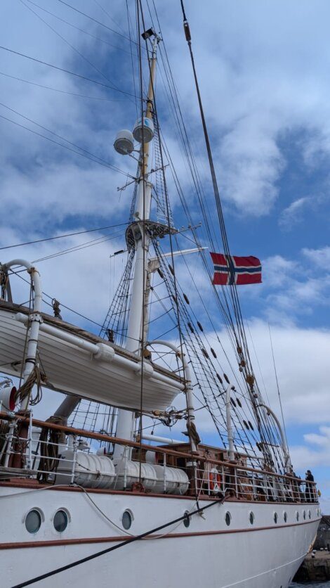 A white sailing ship with a Norwegian flag is moored under a partly cloudy sky, with ropes, lifeboats, and rigging visible.