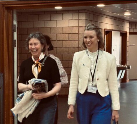 Two women walk together in a corridor, both smiling. One holds a folded blanket, and both wear name badges and lanyards. The background shows brick walls and corridor lighting.