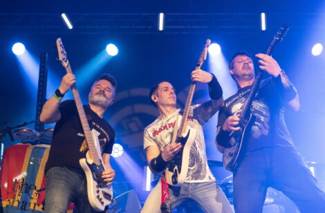 Three guitarists perform energetically on stage under blue and yellow lights, each playing an electric guitar and wearing casual clothes.