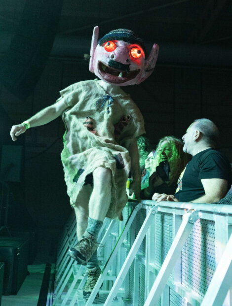 Person wearing a large, glowing red-eyed mask and a tattered costume stands on a barrier at an indoor event, with people watching from behind the rail.