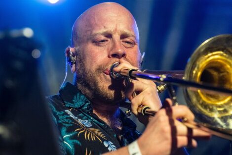 A man with a patterned shirt plays a trombone on stage, wearing an in-ear monitor, with blue stage lighting in the background.