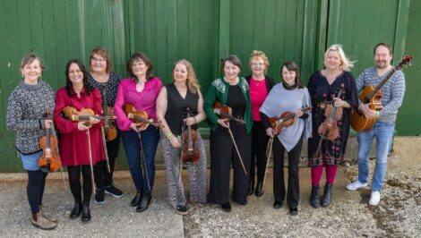 Ten people stand in a row outdoors against a green wooden wall, holding string instruments and smiling at the camera.