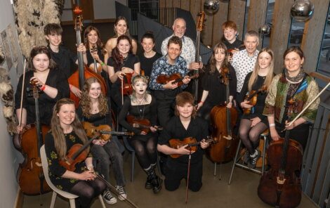 A group of musicians posing indoors with various string instruments, including violins, violas, and cellos, arranged in two rows and smiling at the camera.