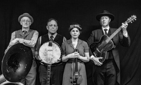 Four musicians in vintage attire stand against a black background, each holding an instrument: a sousaphone, banjo, violin, and guitar.