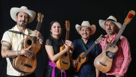Four people wearing hats and traditional clothing stand side by side against a black background, each holding a stringed instrument.