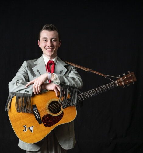 A young man in a grey suit with fringe stands against a black background, smiling and holding an acoustic guitar with the initials "JV" on it.