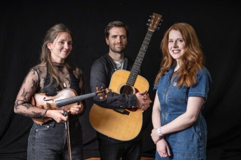 Three people pose in front of a black background; one holds a violin, one holds a guitar, and one stands with hands clasped, all facing the camera.