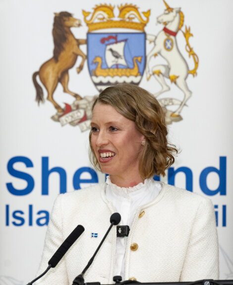 A woman in a white jacket speaks at a lectern in front of a Shetland Islands Council sign and coat of arms.