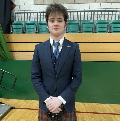 A young person in a navy suit jacket, waistcoat, tie, and tartan kilt stands indoors on a gymnasium floor with folded hands, against rows of green seats and a brick wall.