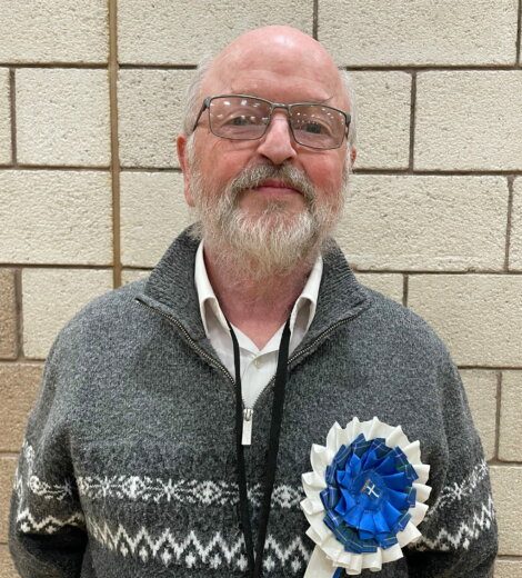An older man with glasses and a grey patterned jumper stands in front of a tiled wall, wearing a large blue and white rosette pinned to his chest.