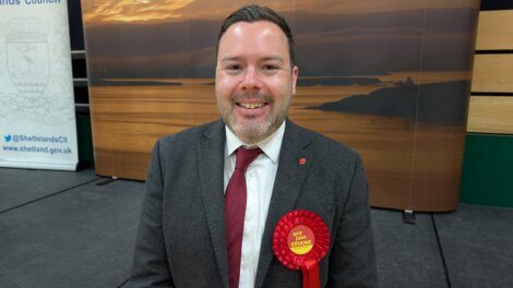 A man in a suit and tie, wearing a red "Labour" rosette, stands indoors in front of a sunset backdrop and Shetland Council banners.