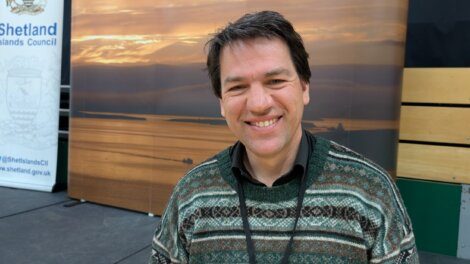 A man in a patterned jumper smiles at the camera, standing indoors near a sunset backdrop and a Shetland Islands Council banner.