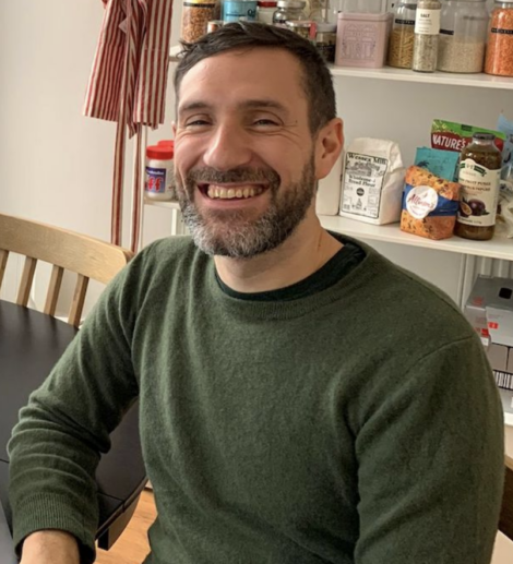 A man with short dark hair and a beard, wearing a green jumper, sits at a table and smiles. Shelves with food items are visible in the background.