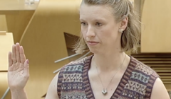 A woman with short blonde hair raises her right hand while being sworn in, wearing a patterned waistcoat and a white rose pinned to her chest.