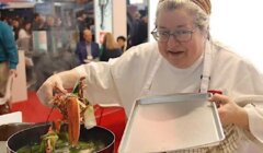 A chef in a white coat lifts a lobster out of a boiling pot with tongs, holding a metal tray in the other hand, as people sit and watch in the background.