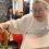 A chef in a white coat lifts a lobster out of a boiling pot with tongs, holding a metal tray in the other hand, as people sit and watch in the background.