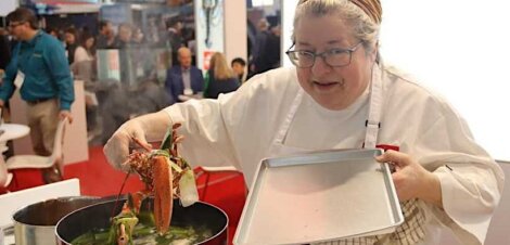 A chef in a white coat lifts a lobster out of a boiling pot with tongs, holding a metal tray in the other hand, as people sit and watch in the background.