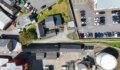 Aerial view of a town area with grey-roofed buildings, a grassy plot, a car park with "One Way" and "No Entry" markings, and white storage