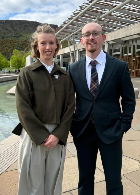 Two people in business attire stand outside a modern building with a pond and greenery in the background on a cloudy day.