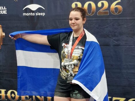A young woman wearing a medal and draped in a blue and white flag holds a trophy in front of a branded backdrop.