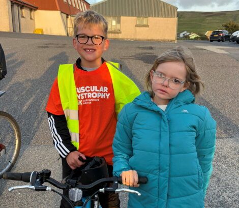 A boy wearing a Muscular Dystrophy orange shirt and high-visibility vest stands with a girl in a teal puffer jacket; both wear glasses and hold bicycle handlebars.