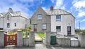 Two-storey semi-detached grey rendered houses with slate roofs under a blue sky, with a low stone wall, red gate and green wheelie bin in