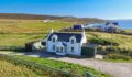 Aerial view of a white two-storey cottage with dark roof, grey outbuildings and a gravel driveway, set on grassy moorland beside a bay with