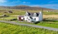 Aerial view of a white-rendered cottage with dormer windows and a slate roof, set beside a stone wall and gravel road, with open moorland